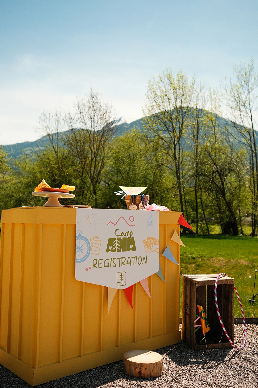 camp-ana-comptoir-accueil-scenographie-enfants Comptoir d’inscription jaune créé pour Camp ANA, avec signalétique personnalisée et guirlandes colorées lors d’un événement en plein air.