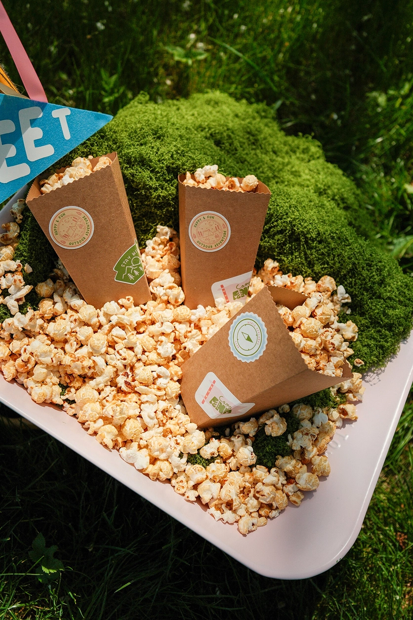 camp-ana-decoration-popcorn-personnalise Cornets de pop-corn personnalisés pour Camp ANA, présentés dans un décor végétal avec des éléments en mousse.