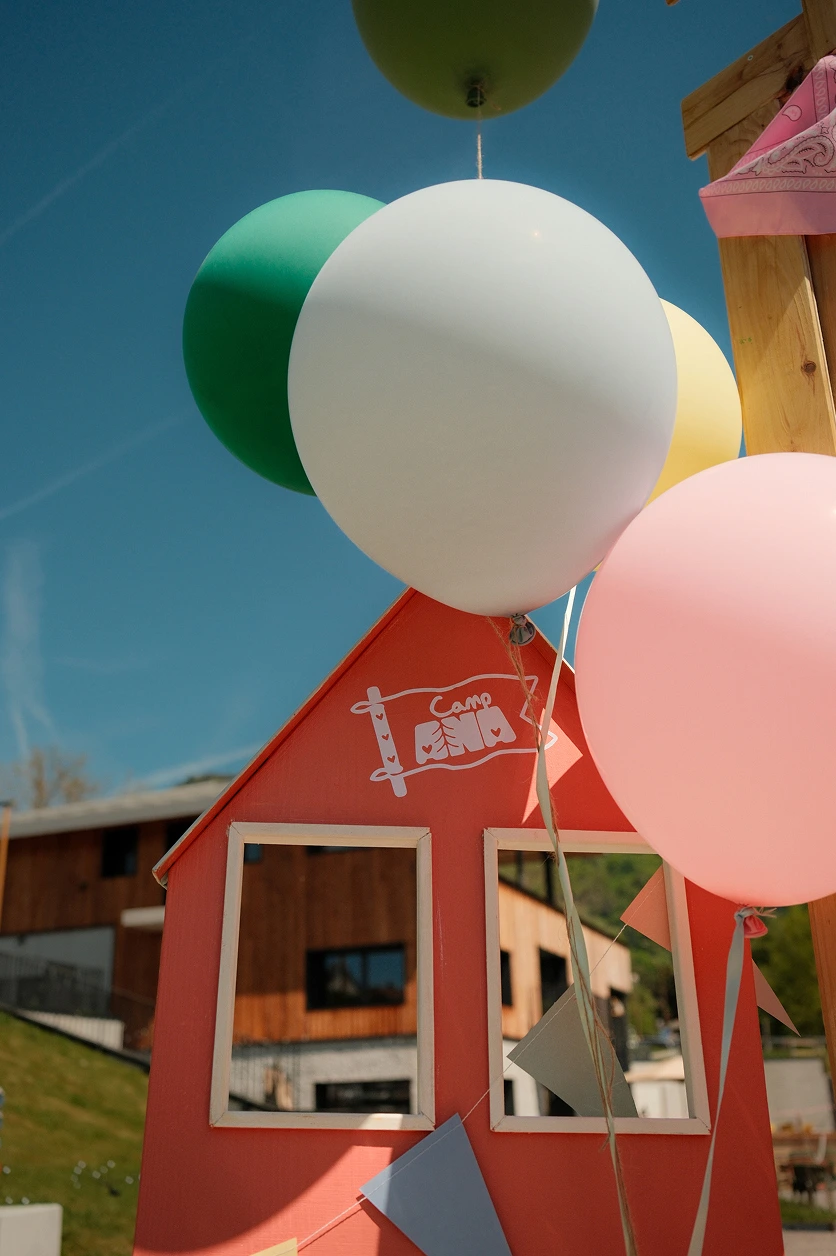 camp-ana-maison-decorative-ballons-pastel Petite maison décorative rose aux couleurs de Camp ANA, accompagnée de ballons pastel et de guirlandes pour un événement familial.