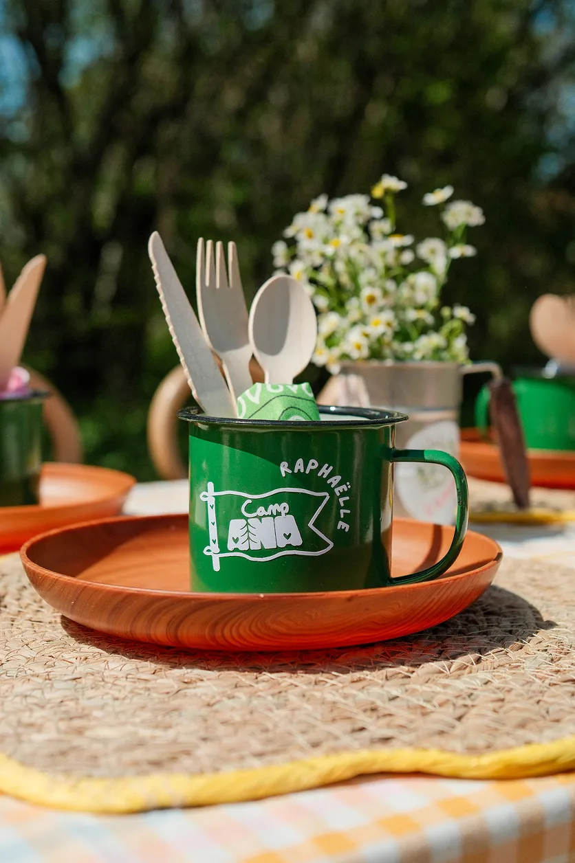 camp-ana-vaisselle-personnalisee-scenographie-enfants-converti-depuis-jpg Mug vert personnalisé Camp ANA avec couverts en bois, bandana et assiette terracotta sur une table décorée pour enfants.