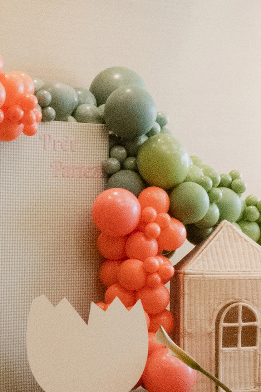 decoration-evenement-enfants-ballons-fleurs-yma Scénographie pour enfants composée d’une arche de ballons verts et corail, d’une petite maison en rotin et de décors floraux.