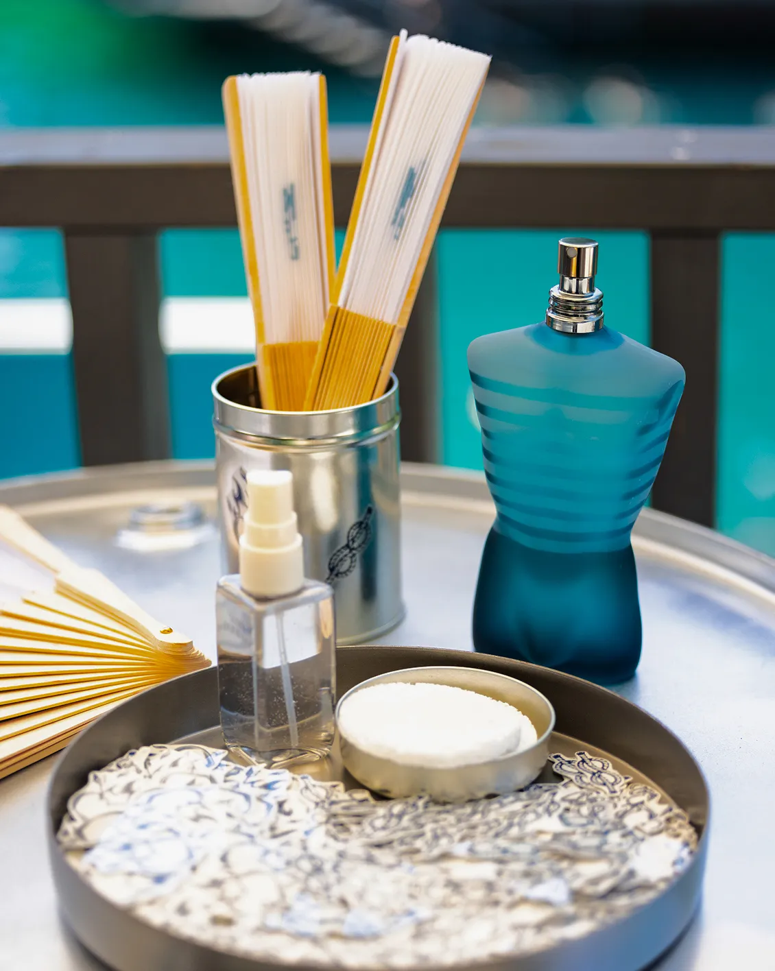 jean-paul-gaultier-lake-club-decoration-personnalisee-converti-depuis-jpg Mise en scène d’un parfum Jean Paul Gaultier sur un plateau métallique, accompagné d’éventails, de flacons et d’accessoires aux tons bleus et blancs.