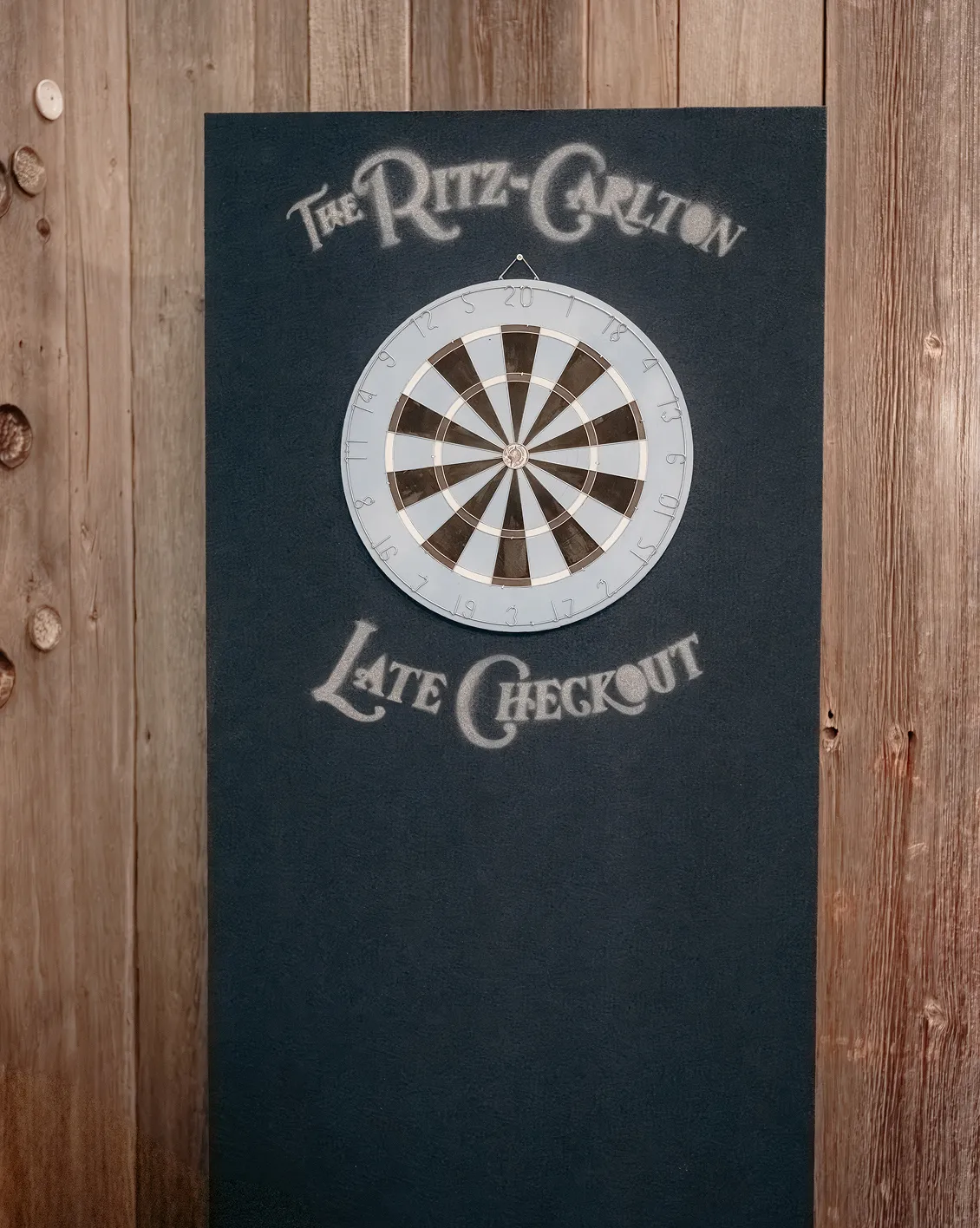 ritz-carlton-late-checkout-animation-jeu-flechettes-converti-depuis-jpg Jeu de fléchettes personnalisé aux couleurs du Ritz-Carlton Late Checkout, installé sur un panneau bleu foncé devant un mur en bois.