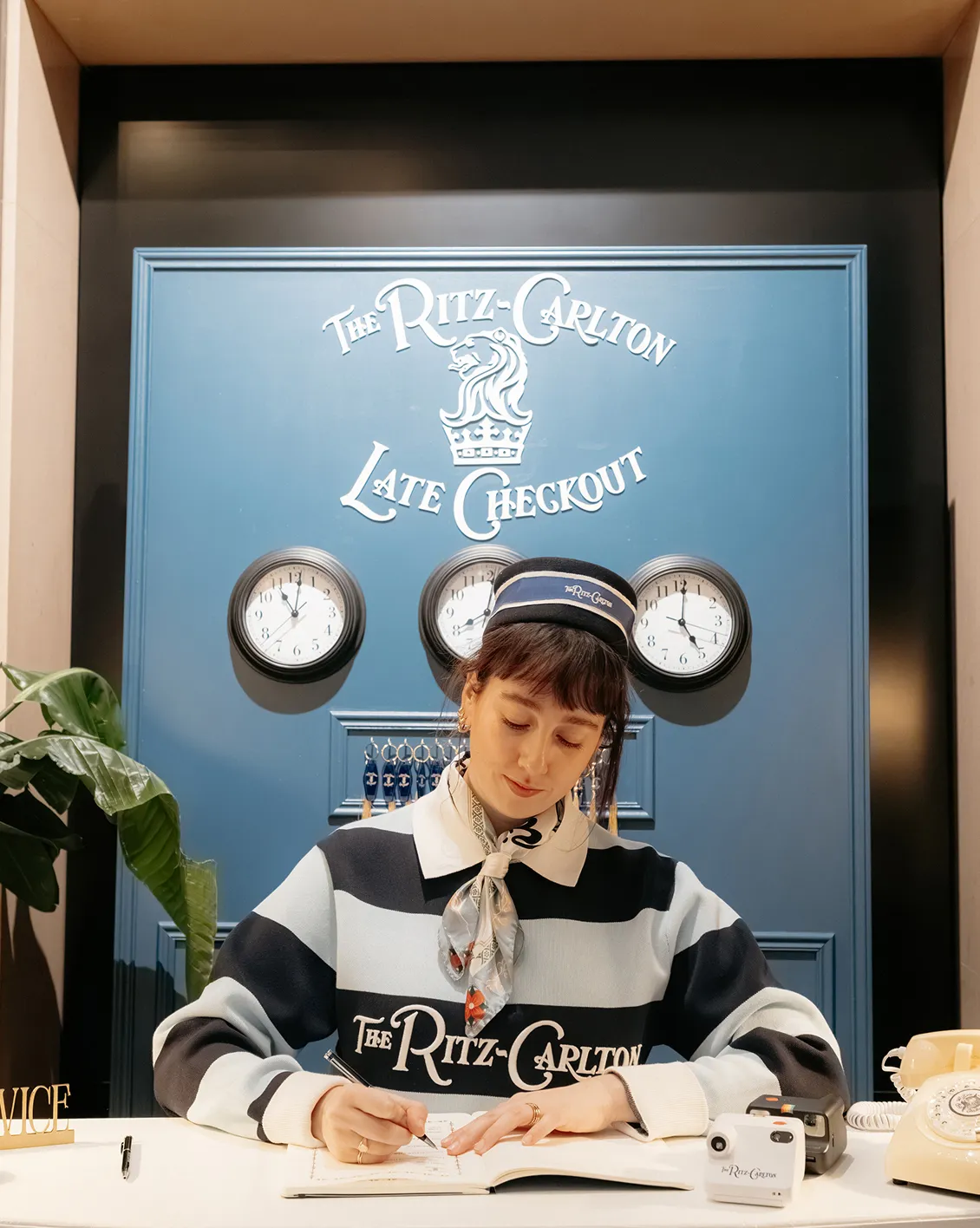 ritz-carlton-late-checkout-scenographie-reception-converti-depuis-jpg Scénographie événementielle The Ritz-Carlton Late Checkout avec comptoir de réception bleu, horloges murales et hôtesse en uniforme personnalisé.