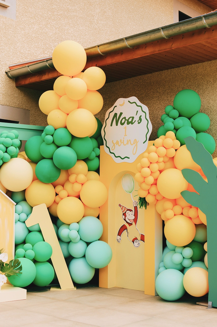scenographie-anniversaire-1-an-noa-yma Scénographie du premier anniversaire de Noa avec une arche décorative, des ballons verts et jaunes et un décor sur le thème de la jungle.