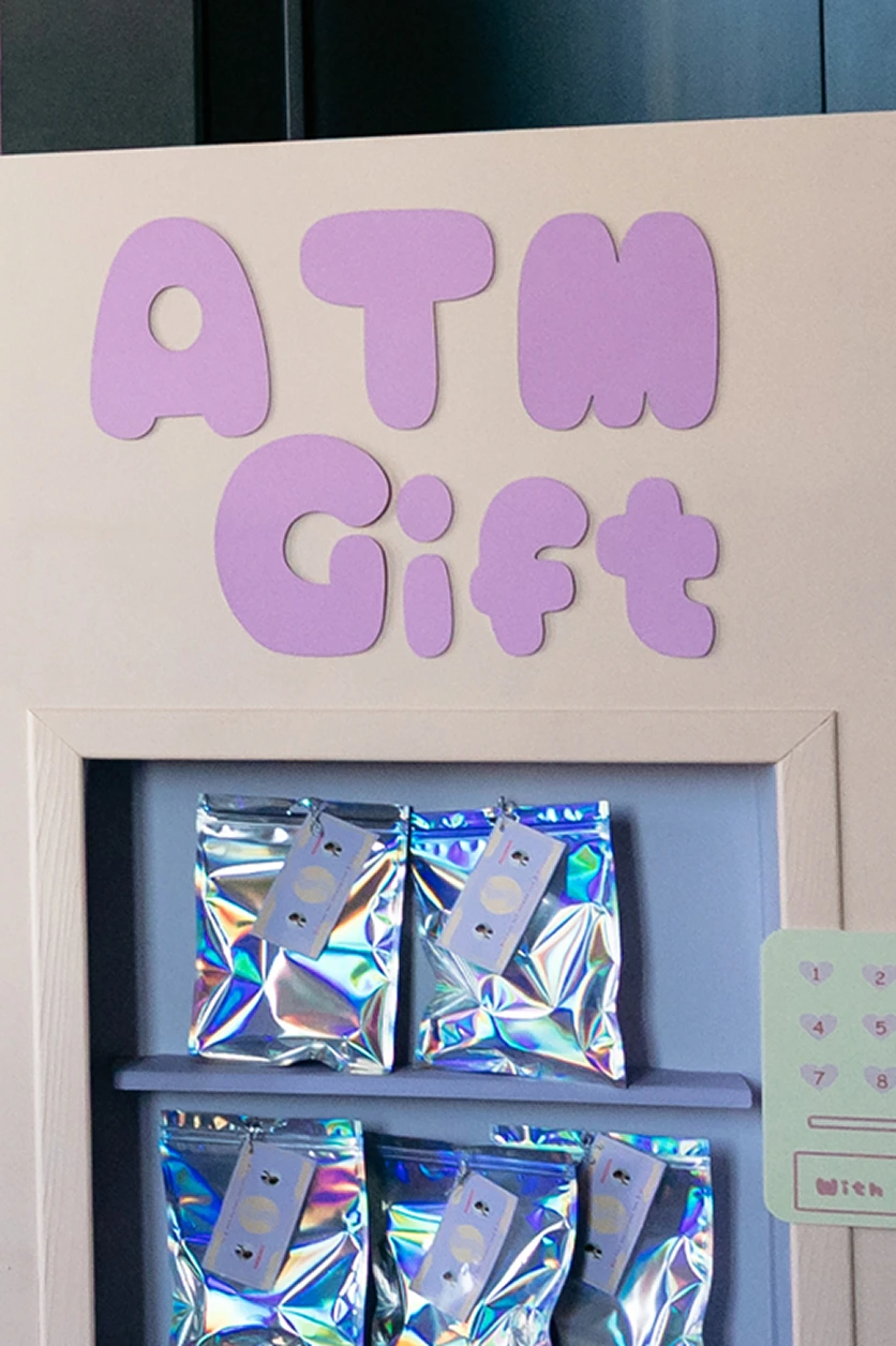 scenographie-espace-cadeaux-atm-gift-yma Espace cadeaux ATM Gift avec enseigne violette et présentoir de pochettes holographiques, dans un décor aux couleurs pastel.